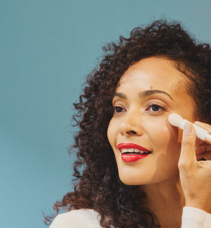 Woman with brown eyes, brown curly hair and red lipstick holding white makeup brush under her eye with a blue background.