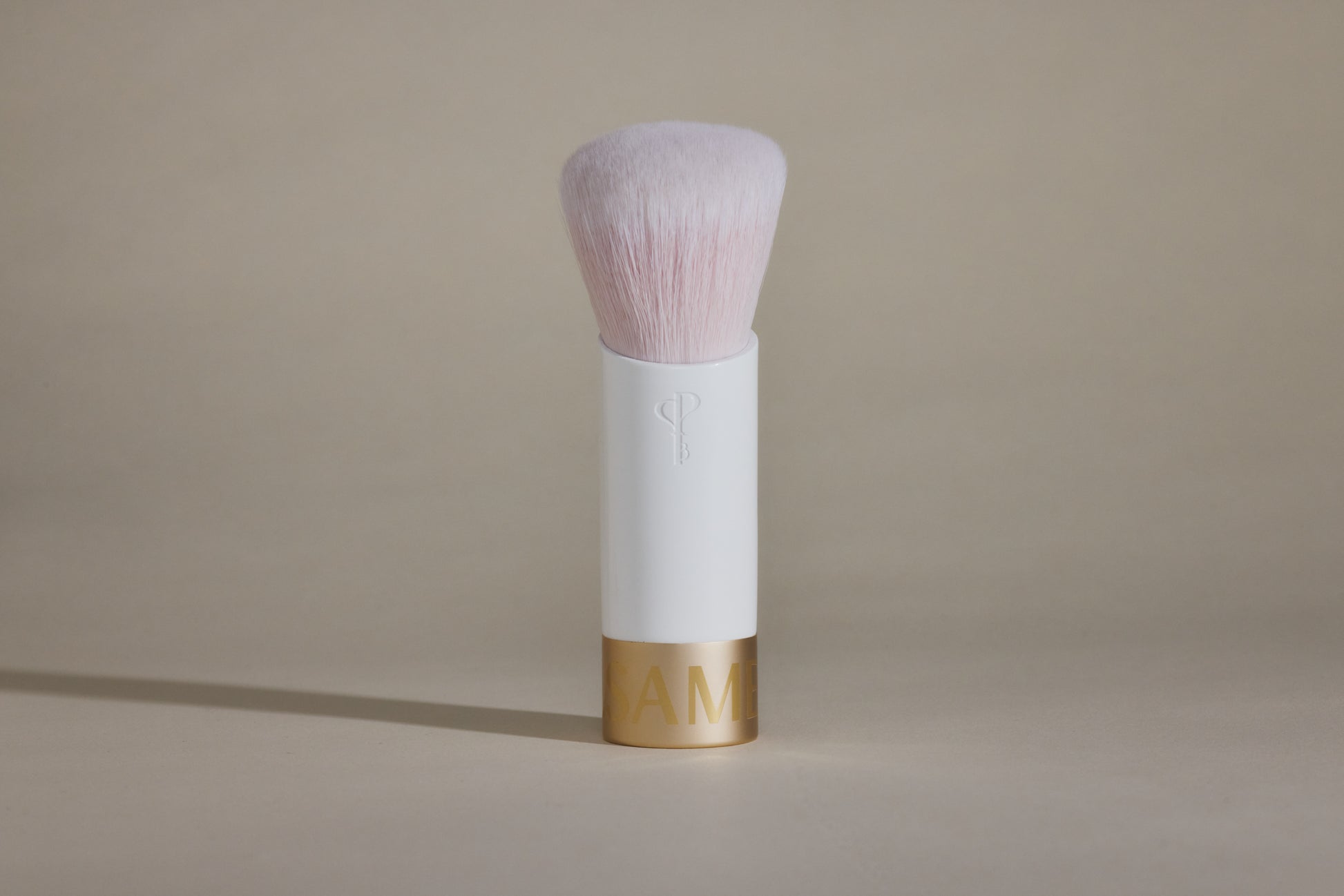 White makeup brush with light pink bristles and gold cap on bottom, standing up on taupe background.