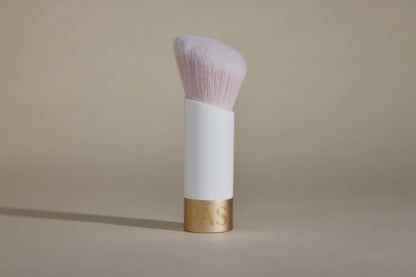 Profile side angle of white makeup brush with light pink bristles and gold cap on bottom, standing up on taupe background.