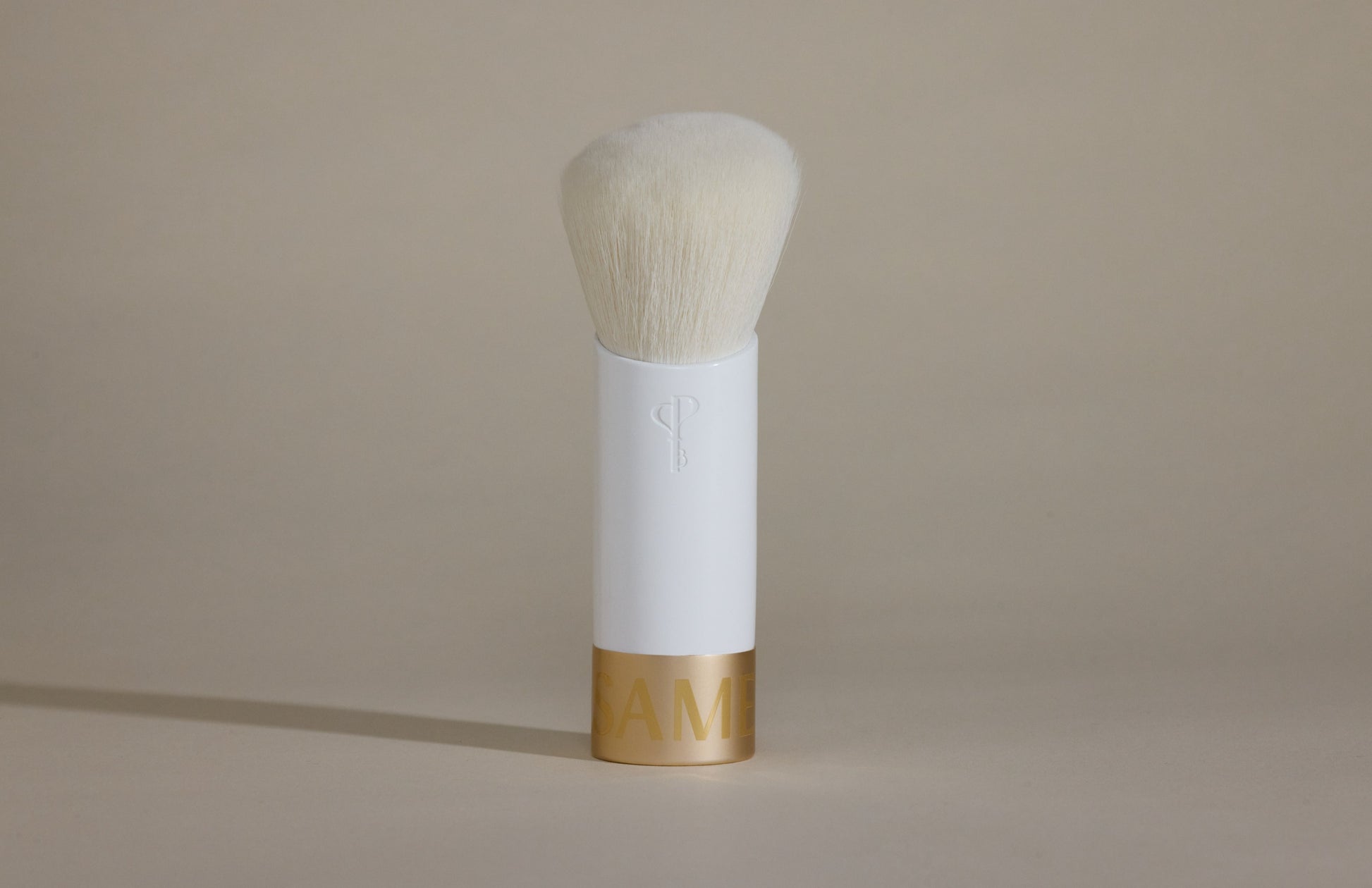 White makeup brush with white bristles and gold cap on bottom, standing up on taupe background.