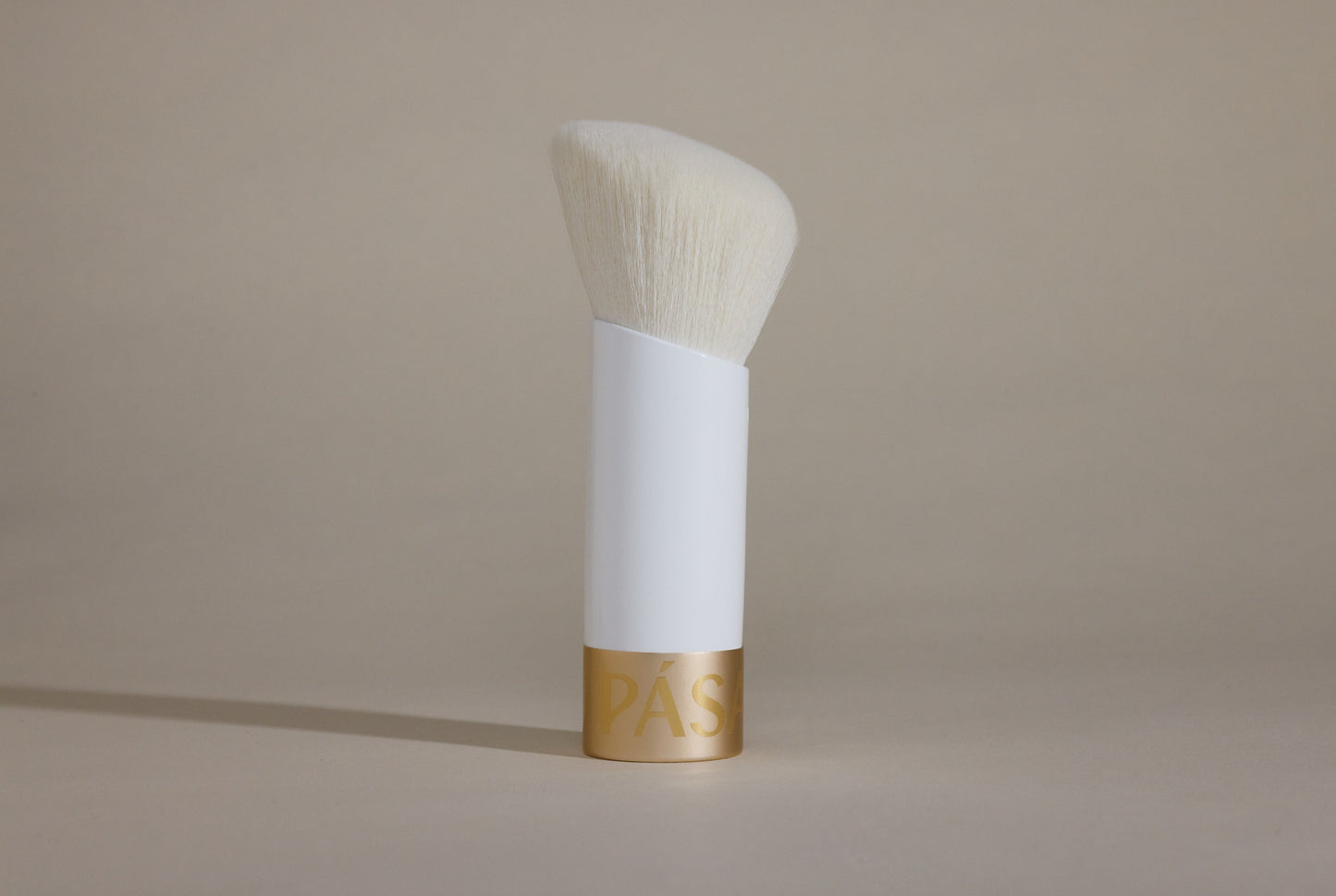 Profile side angle of white makeup brush with white bristles and gold cap on bottom, standing up on taupe background.