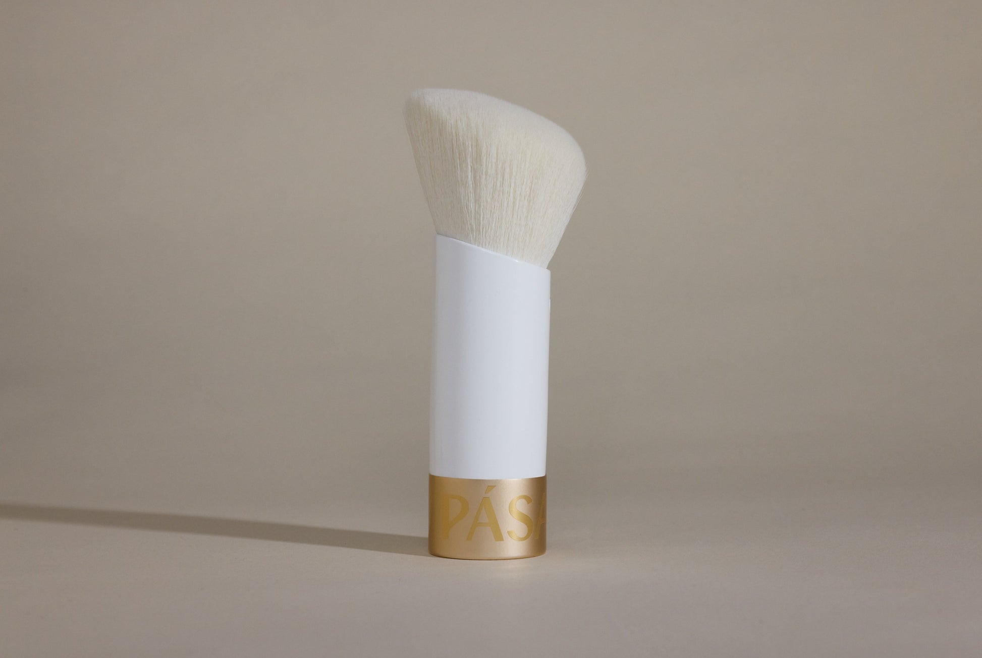 Profile side angle of white makeup brush with white bristles and gold cap on bottom, standing up on taupe background.