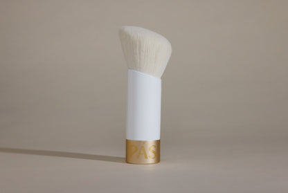 Profile side angle of white makeup brush with white bristles and gold cap on bottom, standing up on taupe background.