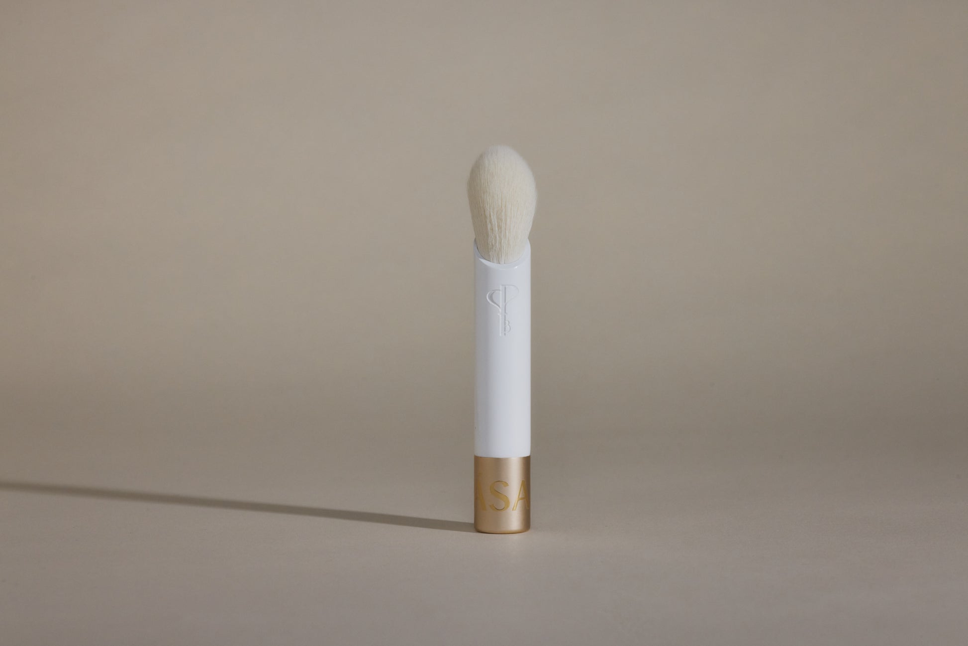 White makeup brush with white bristles and gold cap on bottom, standing up on taupe background.