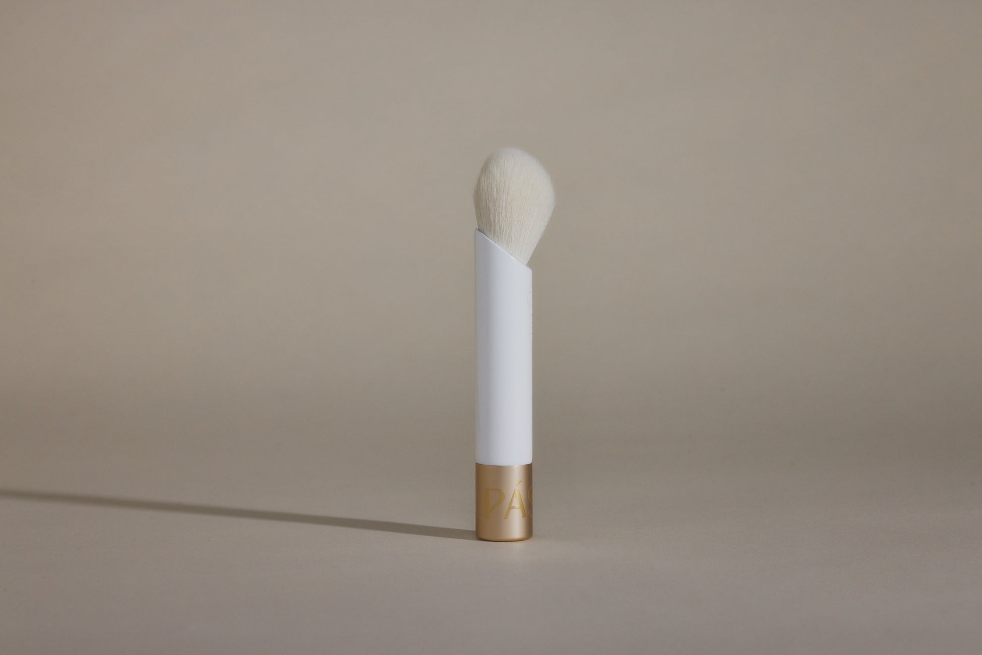 Profile side angle of white makeup brush with white bristles and gold cap on bottom, standing up on taupe background.