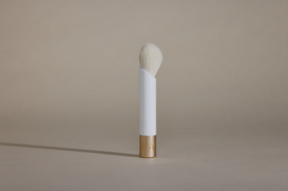 Profile side angle of white makeup brush with white bristles and gold cap on bottom, standing up on taupe background.