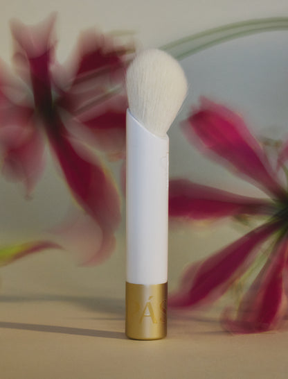 White makeup brush with white bristles and gold cap on bottom pictured with red flowers.
